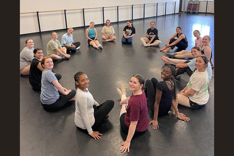 A group of high school dance students sit in a circle and smile for a photo.