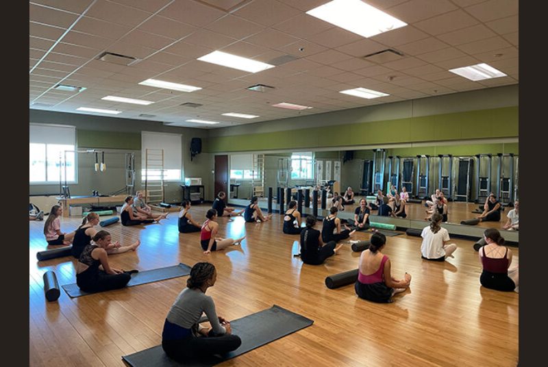 High school dance students sit on the floor of a dance studio, listening to an instructor.