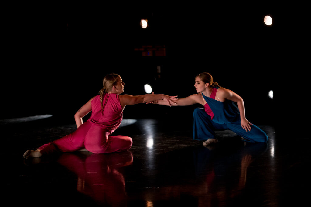 Two dancers are shown on the floor of a black box theater, each reaching out to the other with their right arms.