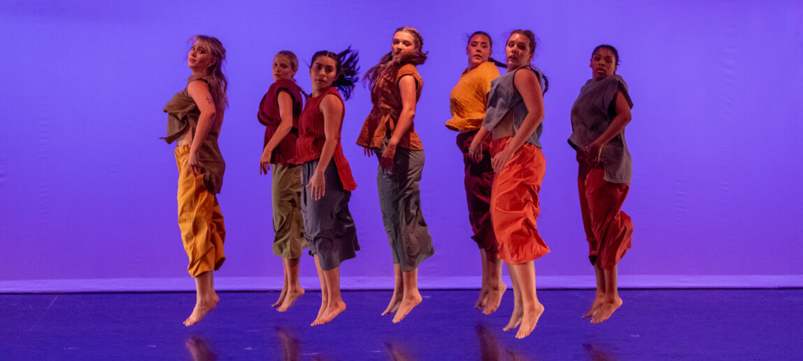 Seven dancers are seen jumping in the air in unison on a stage with a purple-lit background.