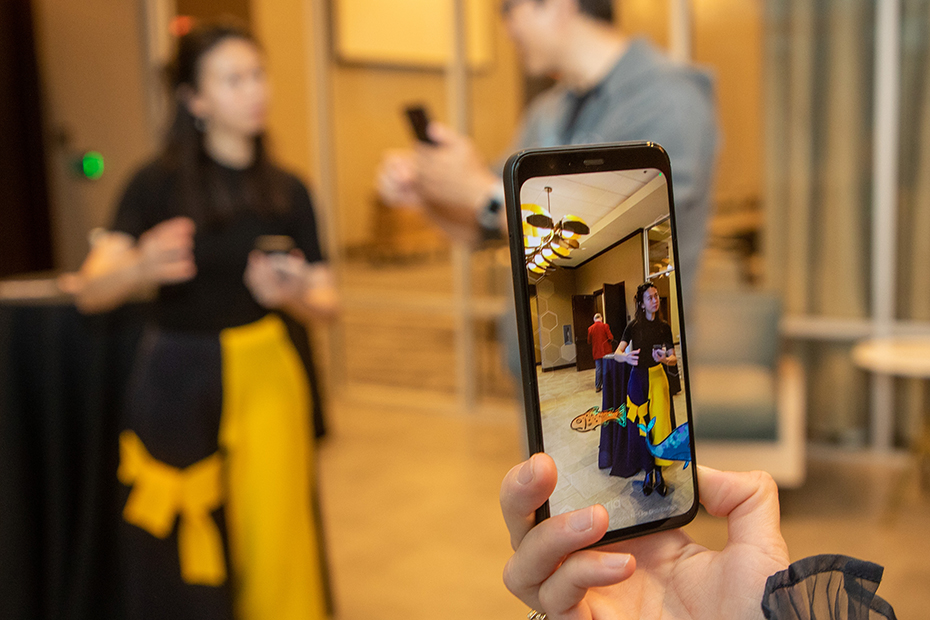 A close-up view of a smartphone that is using an app that incorporates augmented reality. The camera view on the phone shows two people, and a cartoon-like fish is overlaid on top of that view.