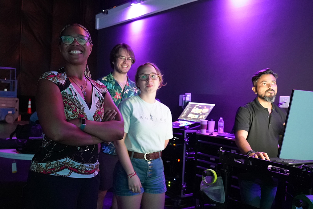 A college professor stands with two students, watching the activity on a virtual production stage (not pictured). To the right is an industry professional in front of a large computer screen.