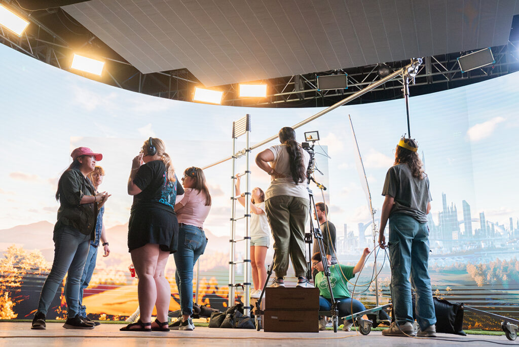 College students work together on a virtual production stage to make a short film. On the large LED screen behind them, a futuristic landscape is shown.