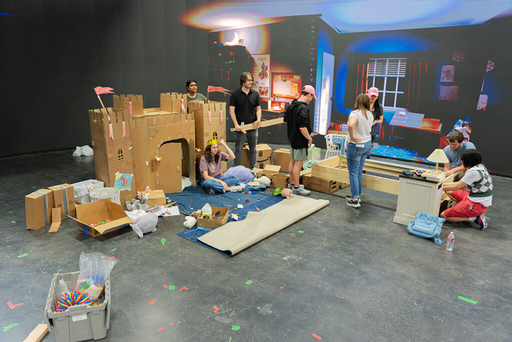 A group of college students work to build a set on a virtual production stage. A cardboard castle is featured, and the image shown on the LED screen is of a child's bedroom.