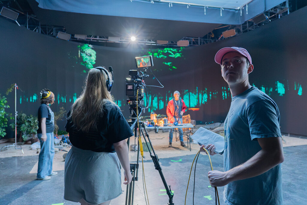 College students work on a scene for a short film on a virtual production stage. An actor is shown in front of table, with a nighttime forest shown on the screen behind him.