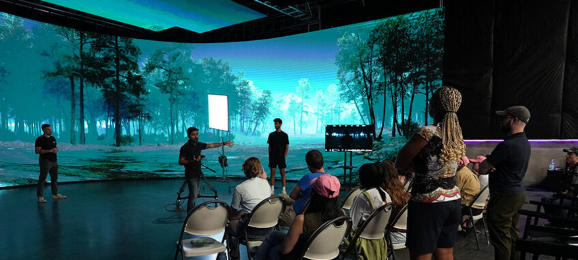 College students and faculty sit and stand on a large virtual production stage, listening to an industry professional speak. On the curved LED wall in front of them, a nighttime forest scene is displayed.