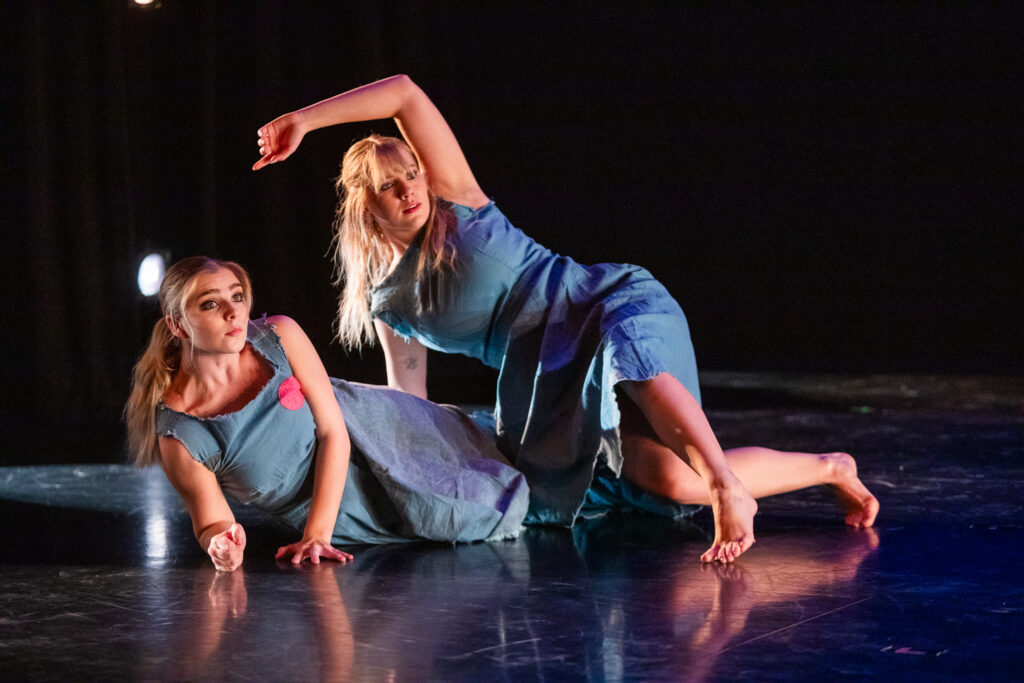 Two dancers, one laying on her side and the other balanced over her, look to the right as they perform onstage.