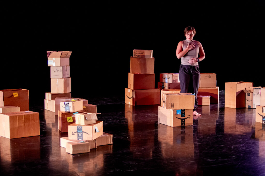 A dancer performs as she is surrounded on a stage by piles of cardboard boxes.