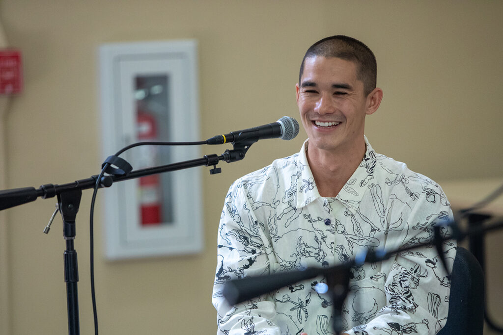 A musician sits in a chair with a microphone in front of him. He smiles as he talks with college students (not pictured).