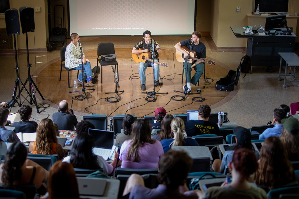 Three musicians sit on a stage in a classroom. Two are holding guitars, all have microphones in front of them. In the foreground is a group of college students, many with laptop computers in front of them, listening to the musicians talk.