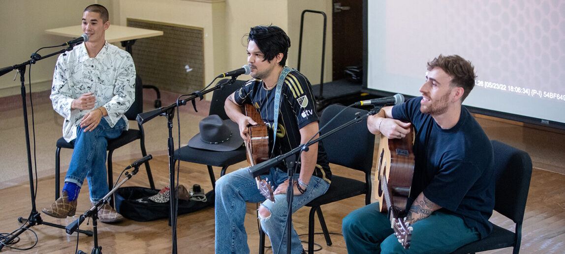 Three musicians sit on a stage in a classroom, speaking with students (not pictured). Two are holding guitars, and all have microphones in front of them.