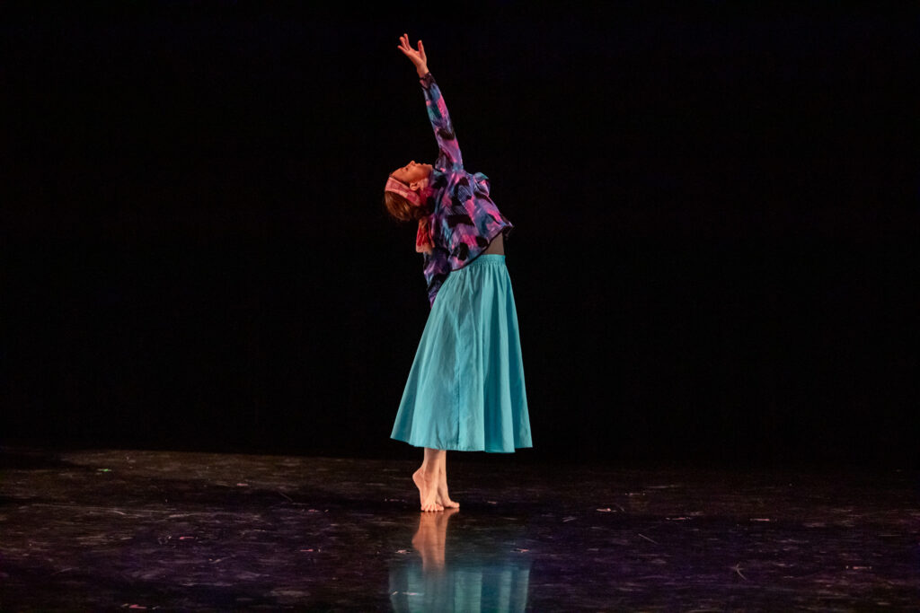 A dancer performs on a darkened stage in a black box theater. She stands up straight on her tiptoes and reaches upward with her right arm.