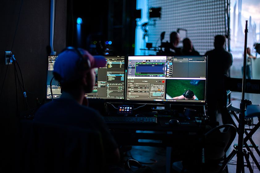 A college student looks at two computer screens displaying information and images from a scene being shot on a virtual production stage in front of him.