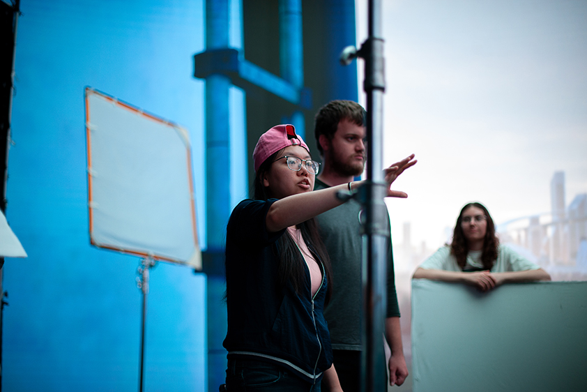 A college student reaches her right arm out as she directs a scene in a short fiml being shot on a virtual production stage. Two college students are listening as she speaks.