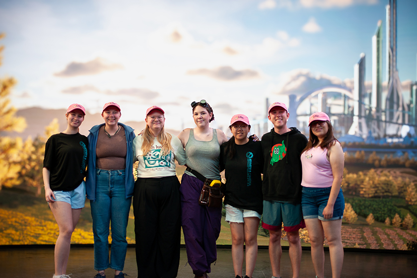 College students smile for a photo with an actor as they stand in front of a large LED screen on a virtual production stage. The screen shows a futuristic landscape.