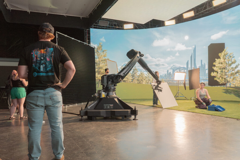 An actor sits in front of an LED screen on a virtual production stage, preparing to shoot a scene for a short film. College students are seen working around the stage.