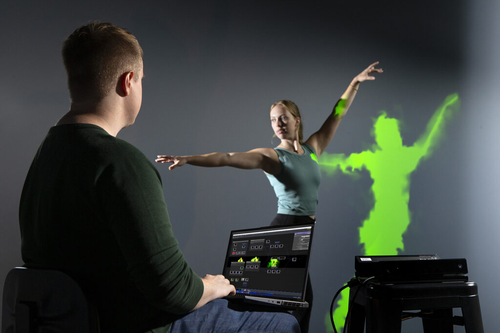 A dancer performs in front of a 360 screen that shows her distorted silhouette in green. In the foreground is a college student working on a laptop computer to create the visual effects.