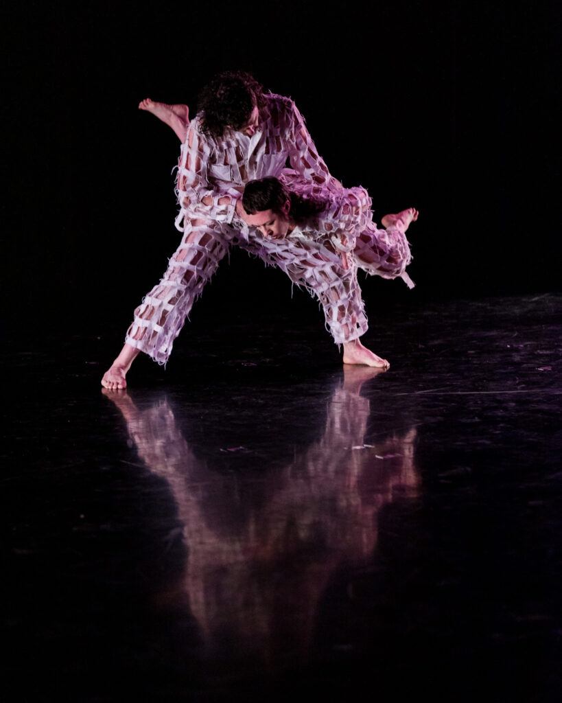 Two dancers perform on a darkened stage in a black box theater, with their reflection visible on the floor below them.