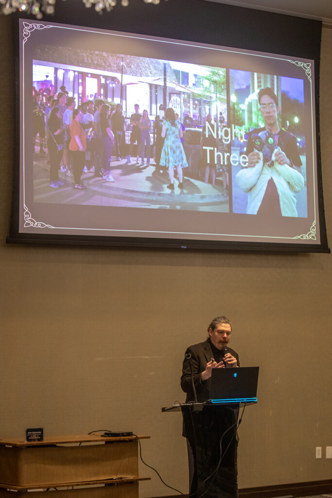 A professor speaks at a podium in a conference room. Projected on the screen above him are two images of a festival.