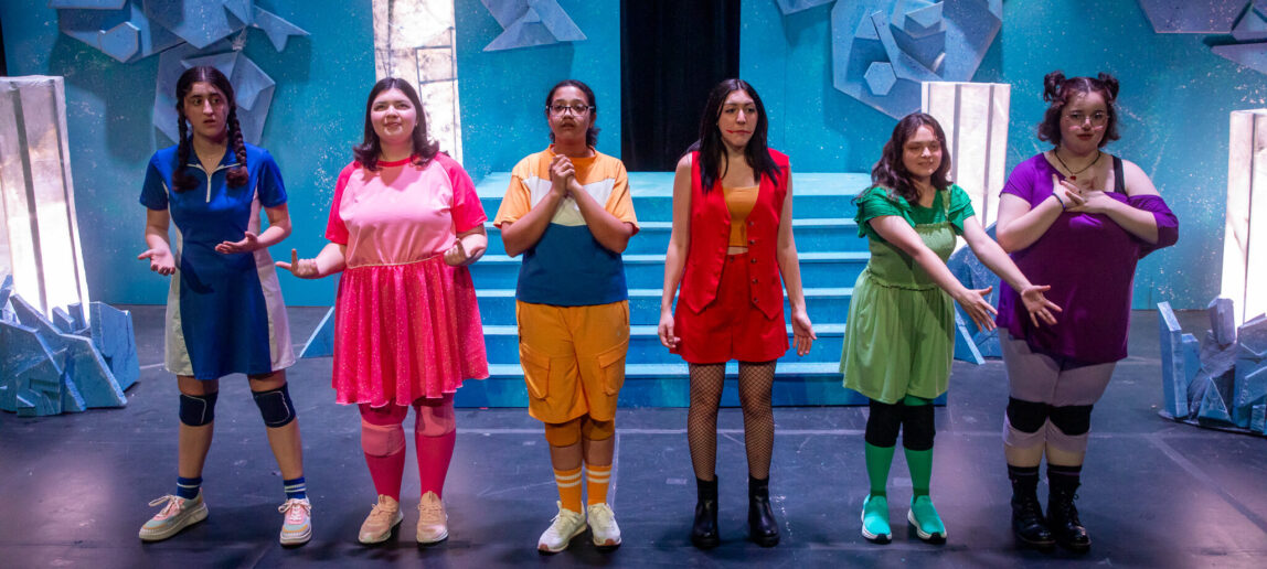 Five student actors, all dressed in a different color (green, pink, blue, purple and orange) perform on stage in a theatrical production.