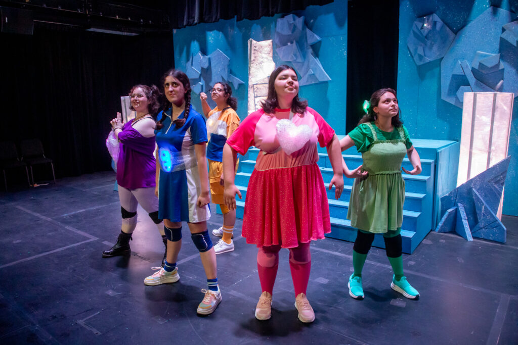 Five student actors, all dressed in a different color (green, pink, blue, purple and orange) perform on stage in a theatrical production.