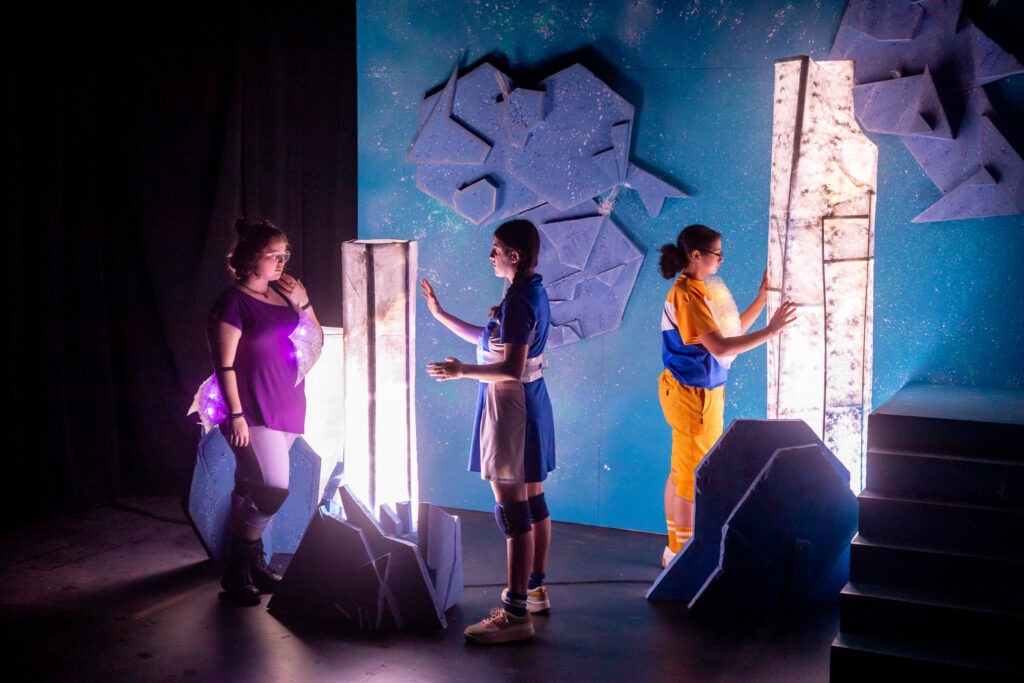 Three student actors interact with lighted column-link props on a stage.