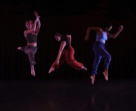 Three dancers perform in a dark studio environment.