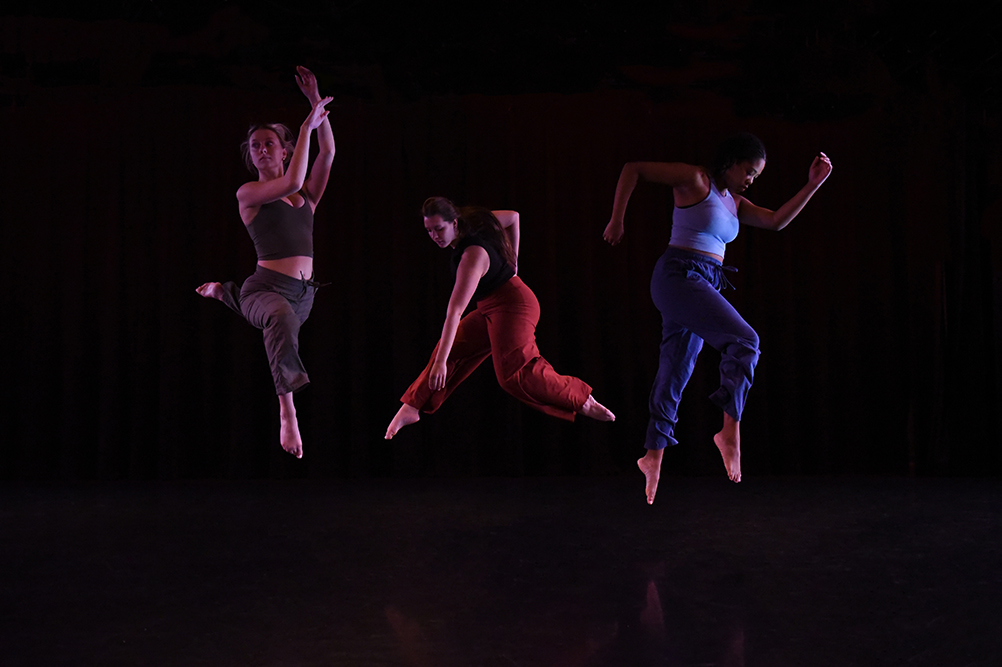 Three dancers perform in a dark studio environment.