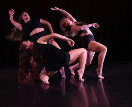 Three dancers perform in a dark studio environment.