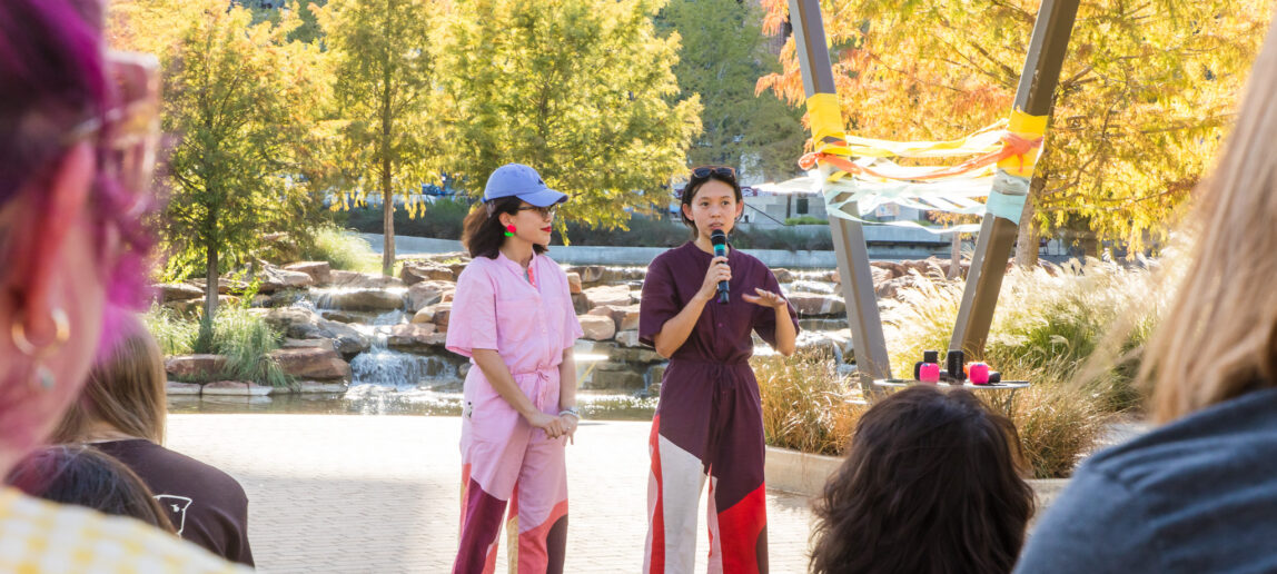 Two women stand on a stage in an outdoor setting. One is speaking into a microphone.