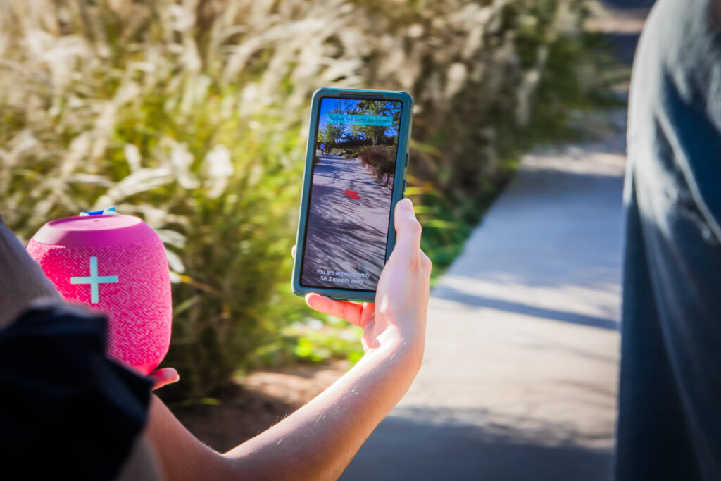 A smartphone is shown with an arrow and message overlaid on the camera view of a path in a park.