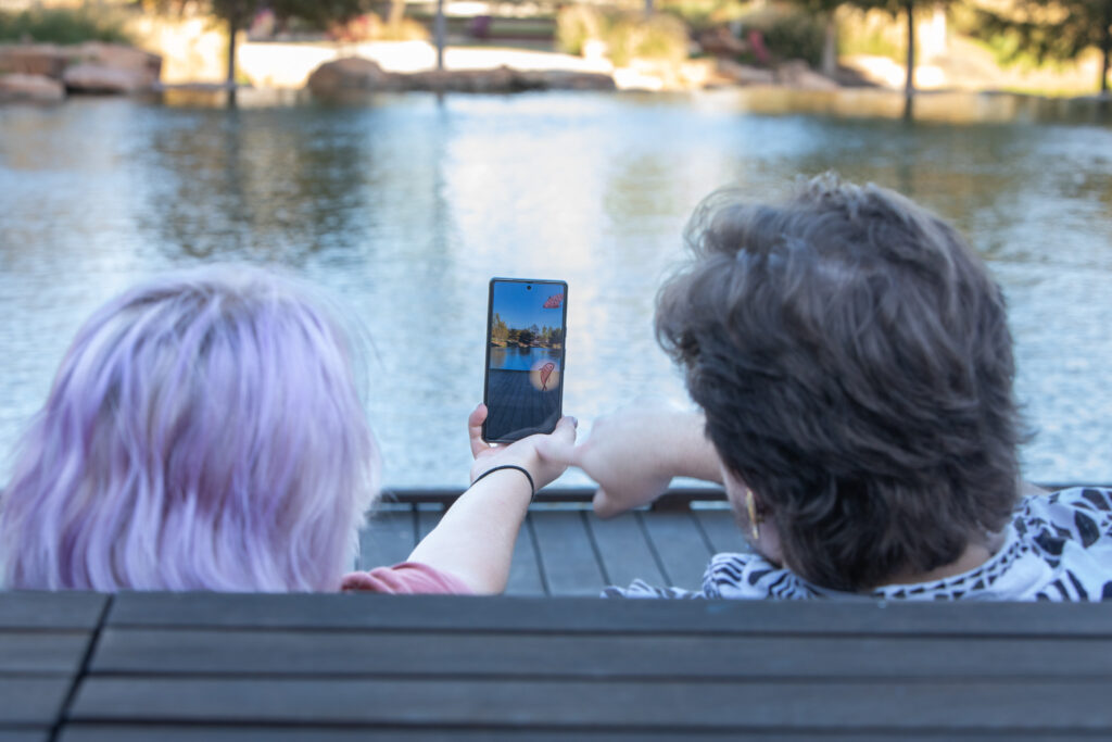 Two people sit in front of a pond. the person on the left is holding a smartphone. Both are looking at the camera view, which shows animated fish characters over the view of the water.