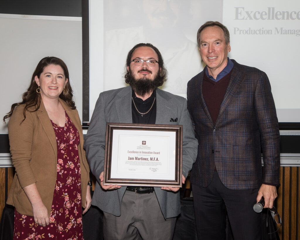 A man holds a framed certificate of achievement. Two university leaders stand alongside him.