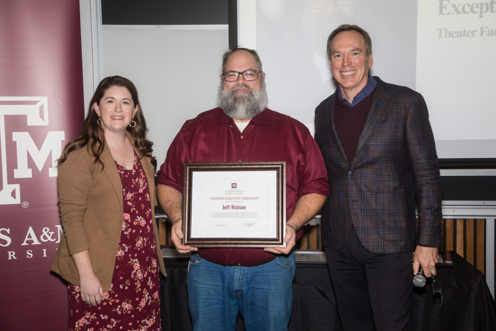 A man holds a framed certificate of achievement. Two university leaders stand alongside him.