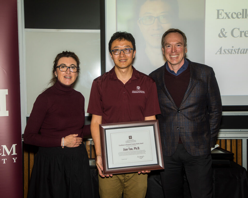 A man holds a framed certificate of achievement. Two university leaders stand alongside him.