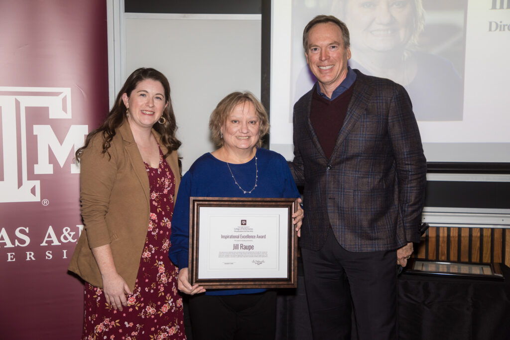 A woman holds a framed certificate of achievement. Two university leaders stand alongside her.