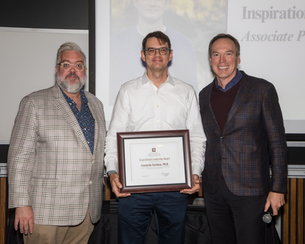 A man holds a framed certificate of achievement. Two university leaders stand alongside him.