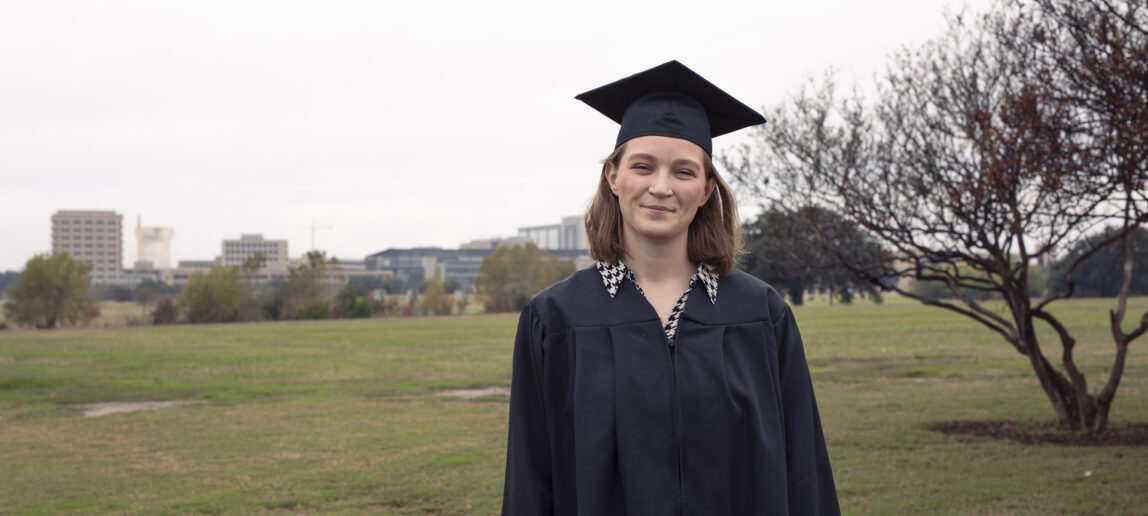 A college student stands on a campus, wearing a traditional cap and gown.