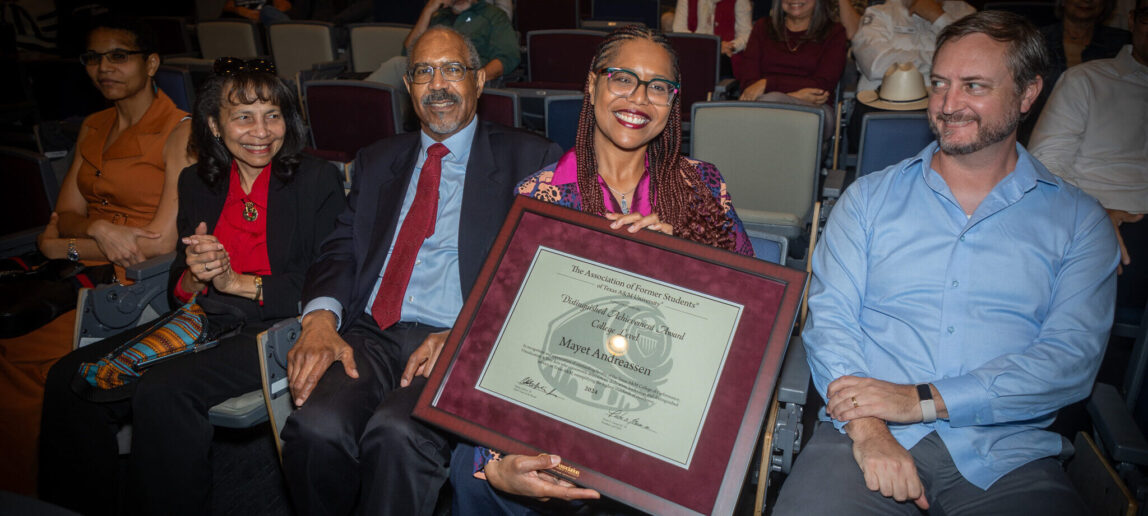 A woman smiles as she holds a framed certificate of recognition, with family members seated around her.