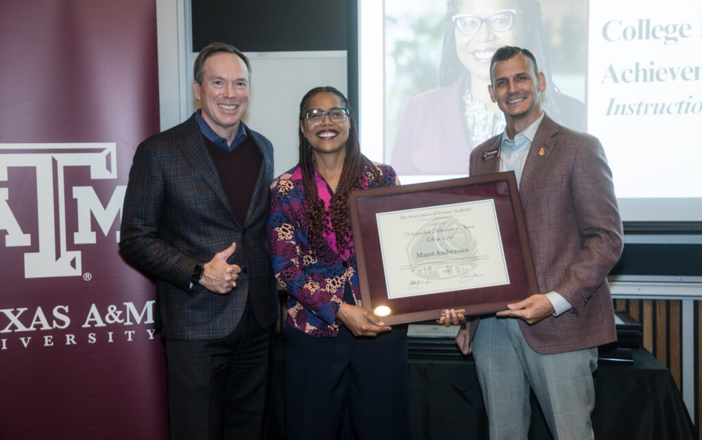 A woman and a man jointly hold a framed certificate of achievement. On their right is a university dean, giving the thumbs-up gesture.