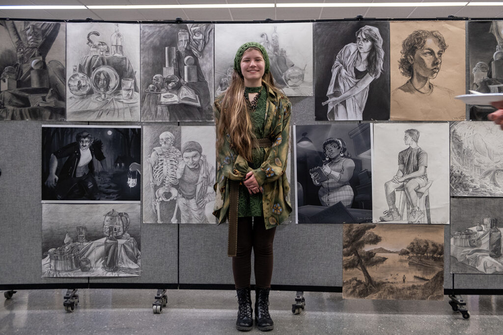 A college student stands in front of a wall that is filled with drawings and sketches.