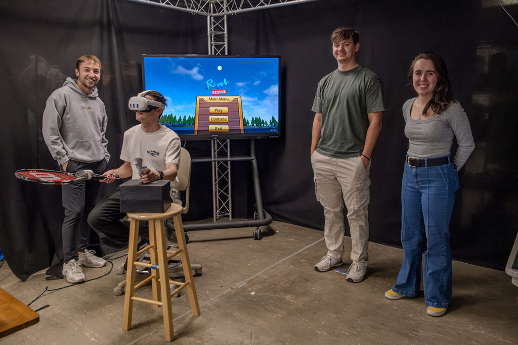 Three college students stand by a monitor showing a virtual reality game. Another student is seated, wearing VR goggles and holding VR devices.