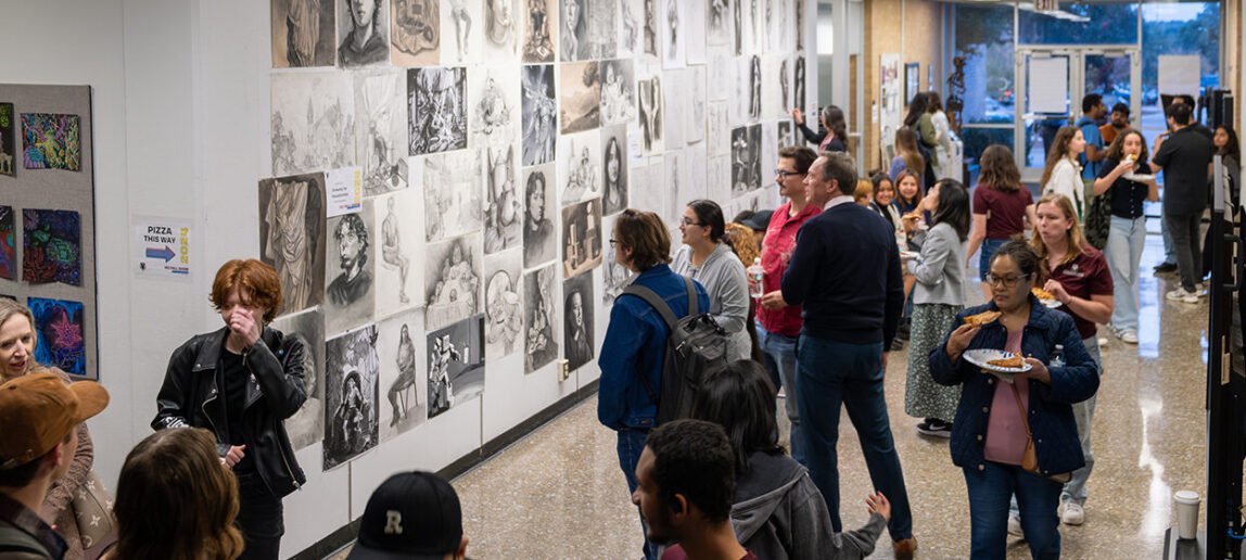 A college hallway is filled with people looking at artwork displayed on the walls.