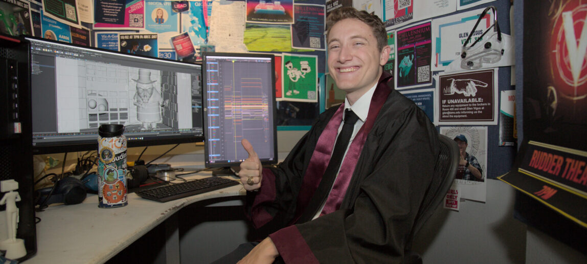 A college student smiles and gives the thumbs-up gesture while sitting at a cubicle with his computer, wearing a graduation gown.