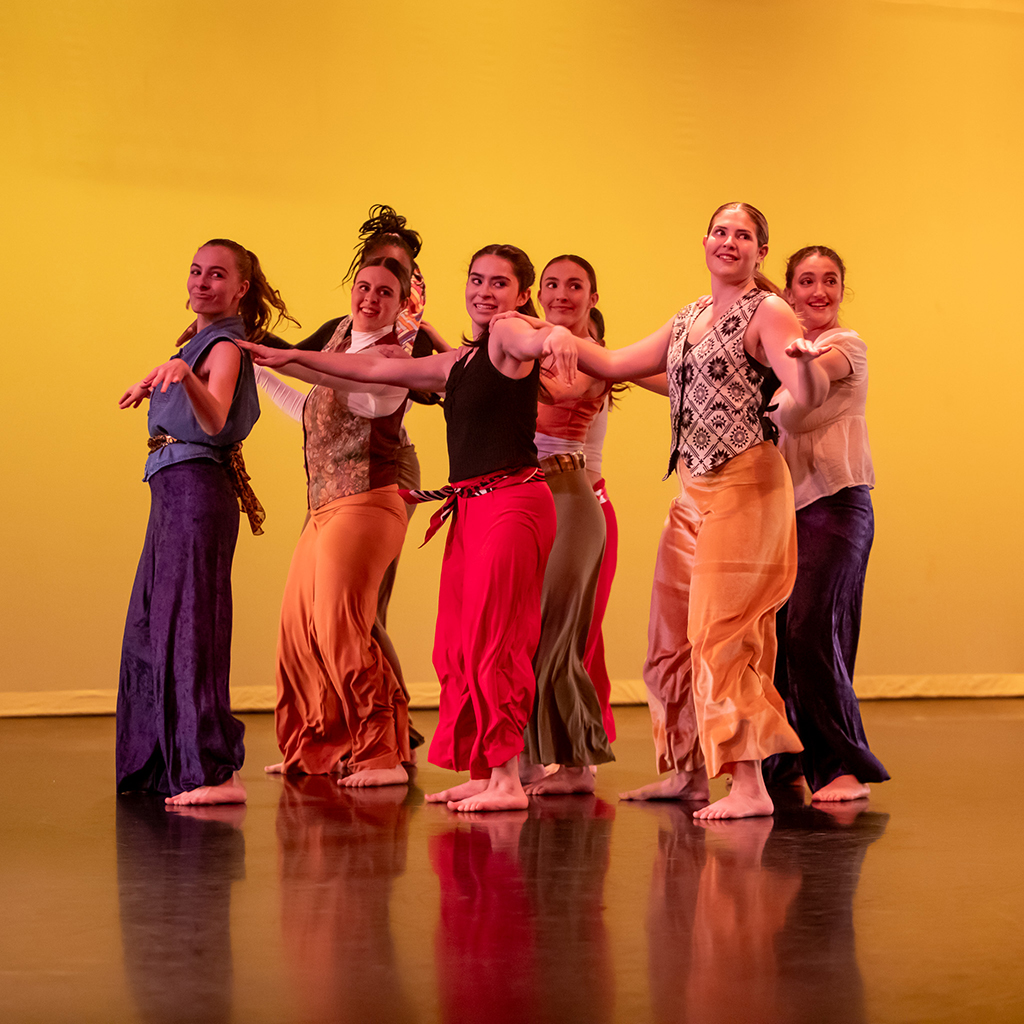 Seven college students wearing colorful outfits perform on stage, with a yellow-lit backdrop behind them.