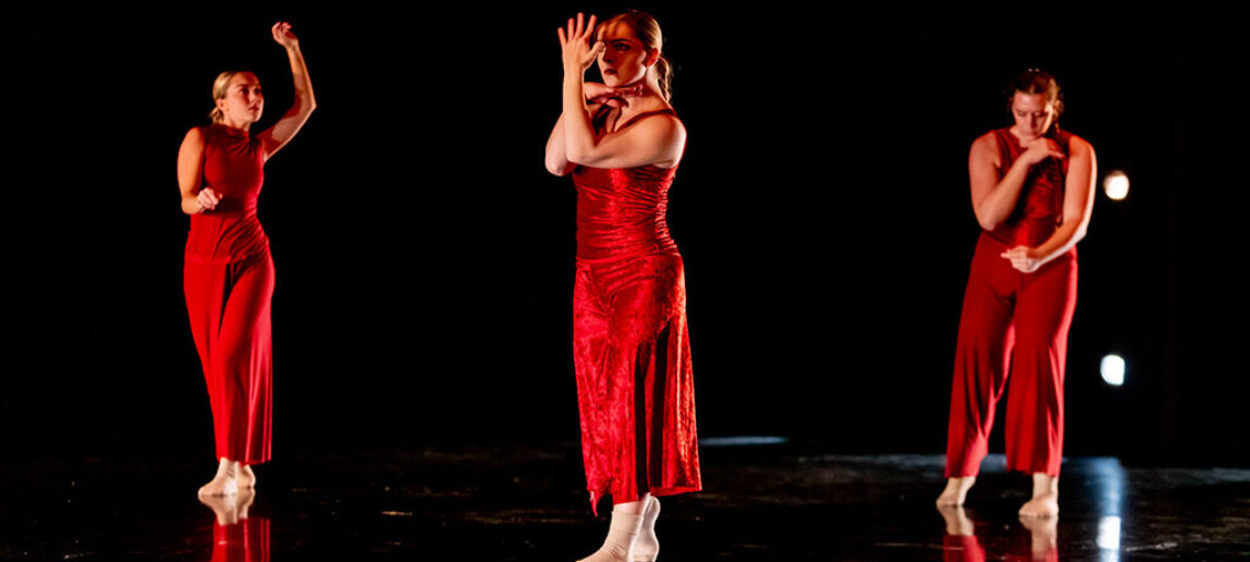 Three college students wearing red outfits perform on a darkened stage.