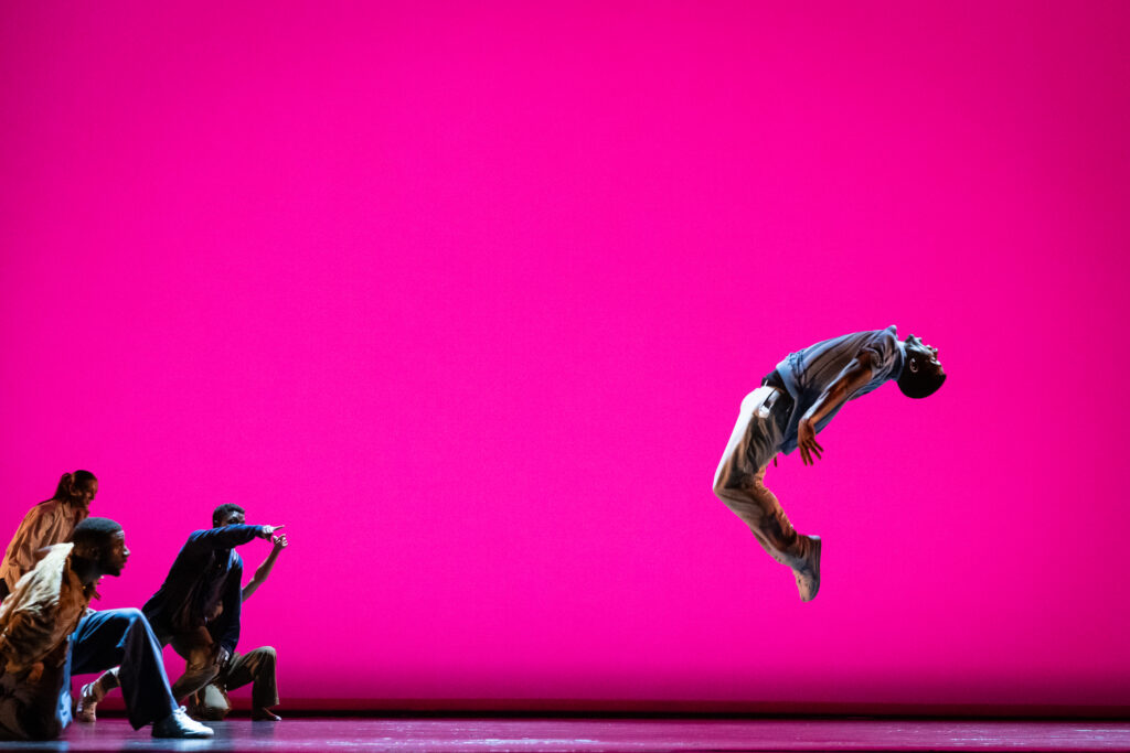 Dancers perform on a stage with a bright pink background. One dancer on the right side is in midair, while several are in the left corner watching.