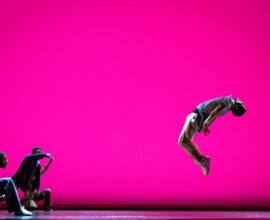 Dancers perform on a stage with a bright pink background. One dancer on the right side is in midair, while several are in the left corner watching.