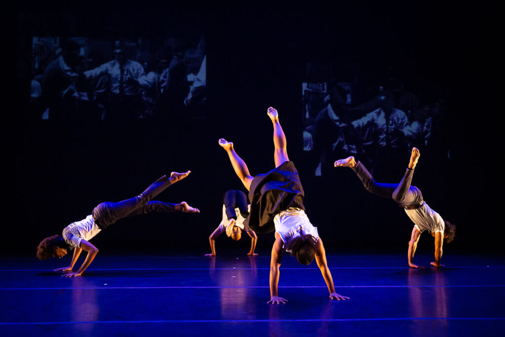 Four dancers have their hands on the stage floor and their bodies and legs extended upward.