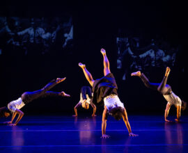 Four dancers have their hands on the stage floor and their bodies and legs extended upward.